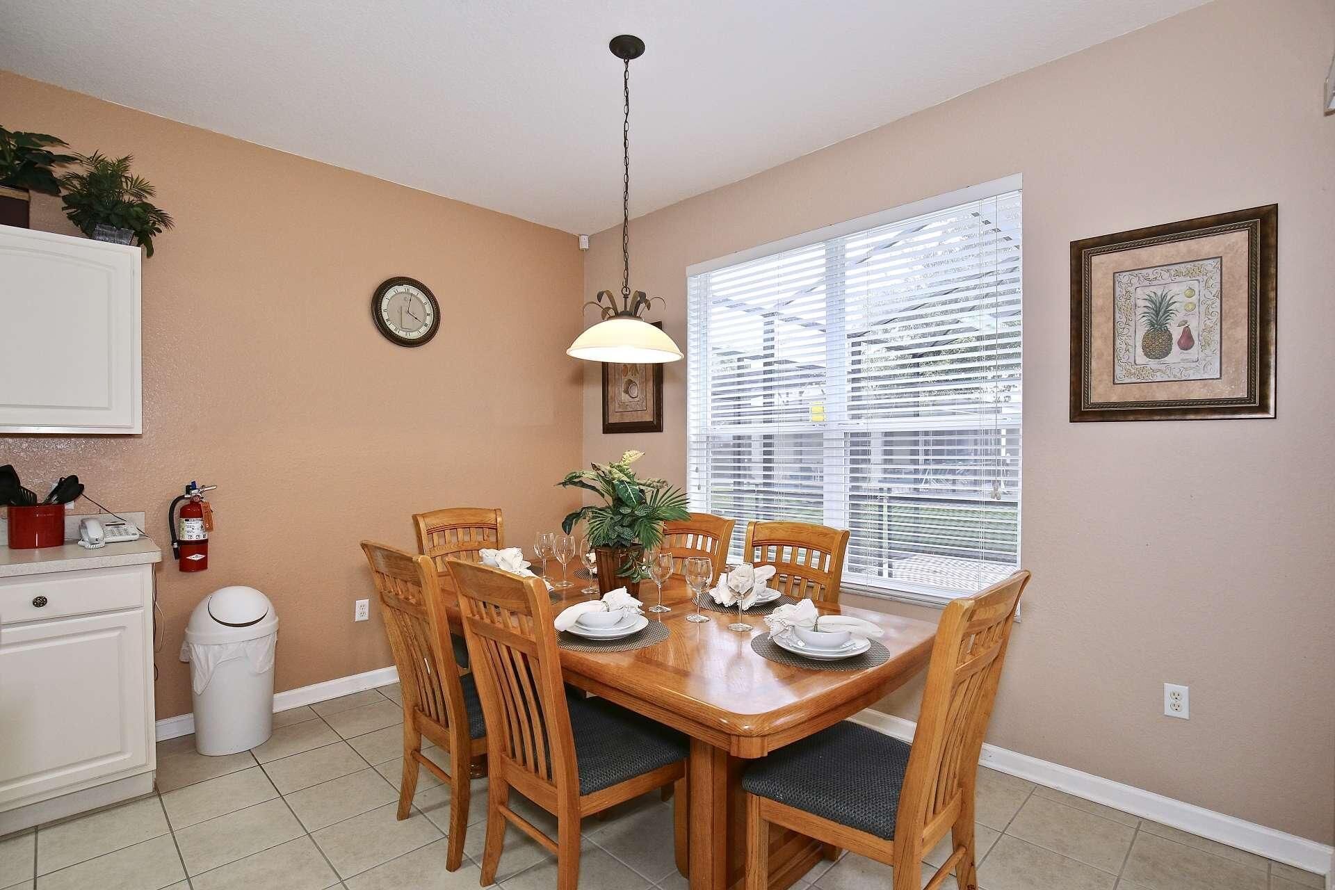 A dining area with a wooden table set for four, surrounded by chairs. There is a clock on the wall, a window with blinds, and a framed picture on the wall.