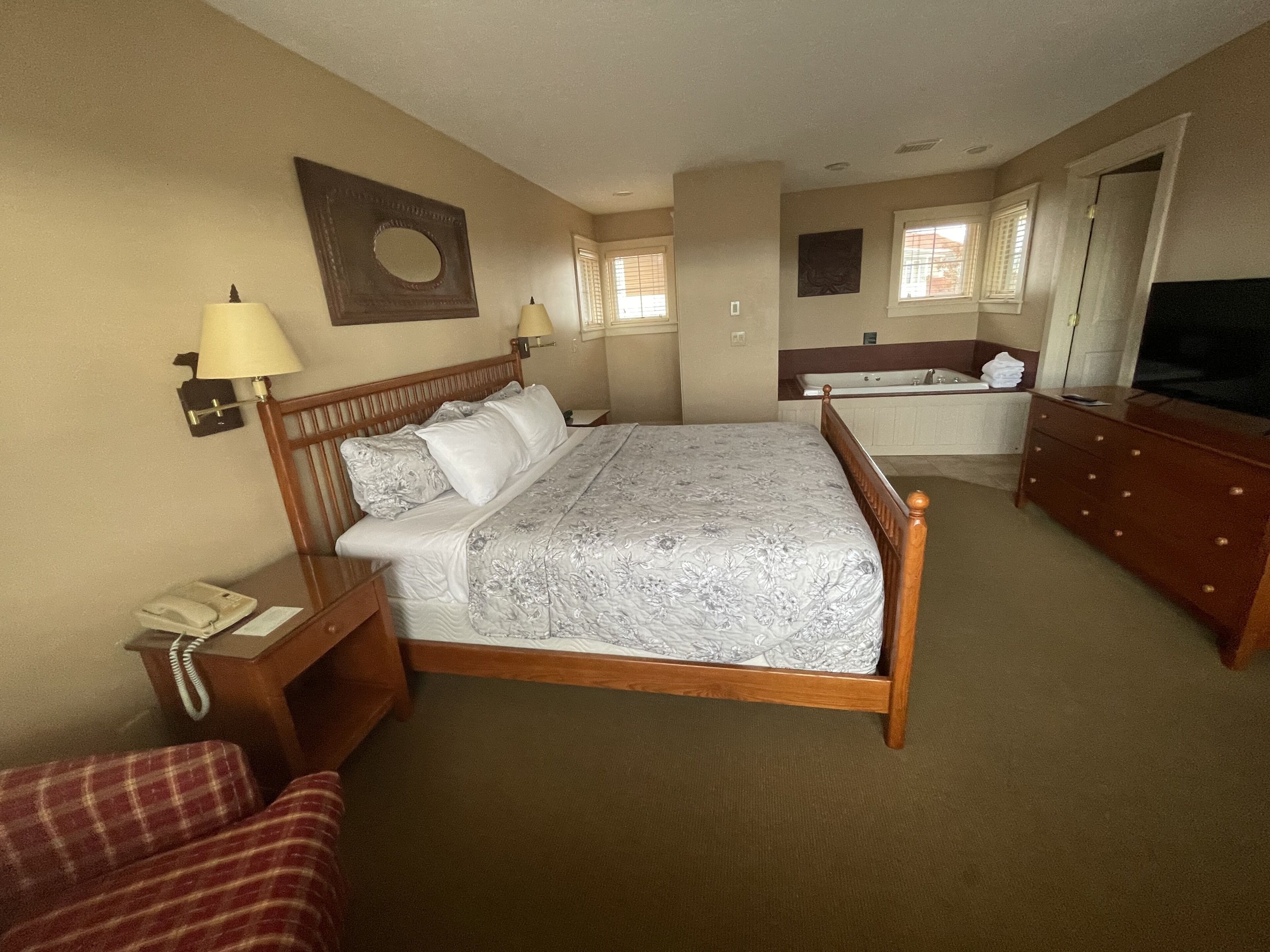A hotel room with a large bed, a wooden dresser, a desk with a lamp, a red plaid chair, a bathroom with a bathtub, and a television.