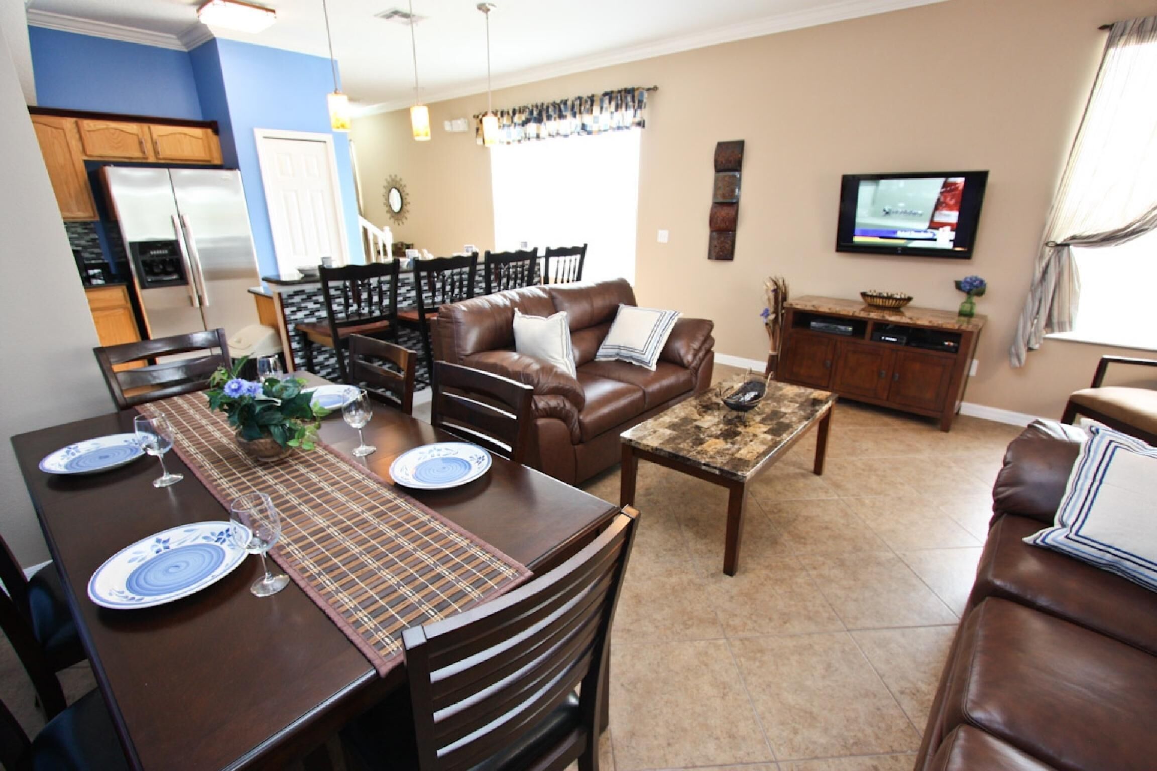 Una sala de estar moderna con un sofá de cuero marrón, una mesa de comedor con platos y vasos, una mesa de centro y un televisor montado en la pared.