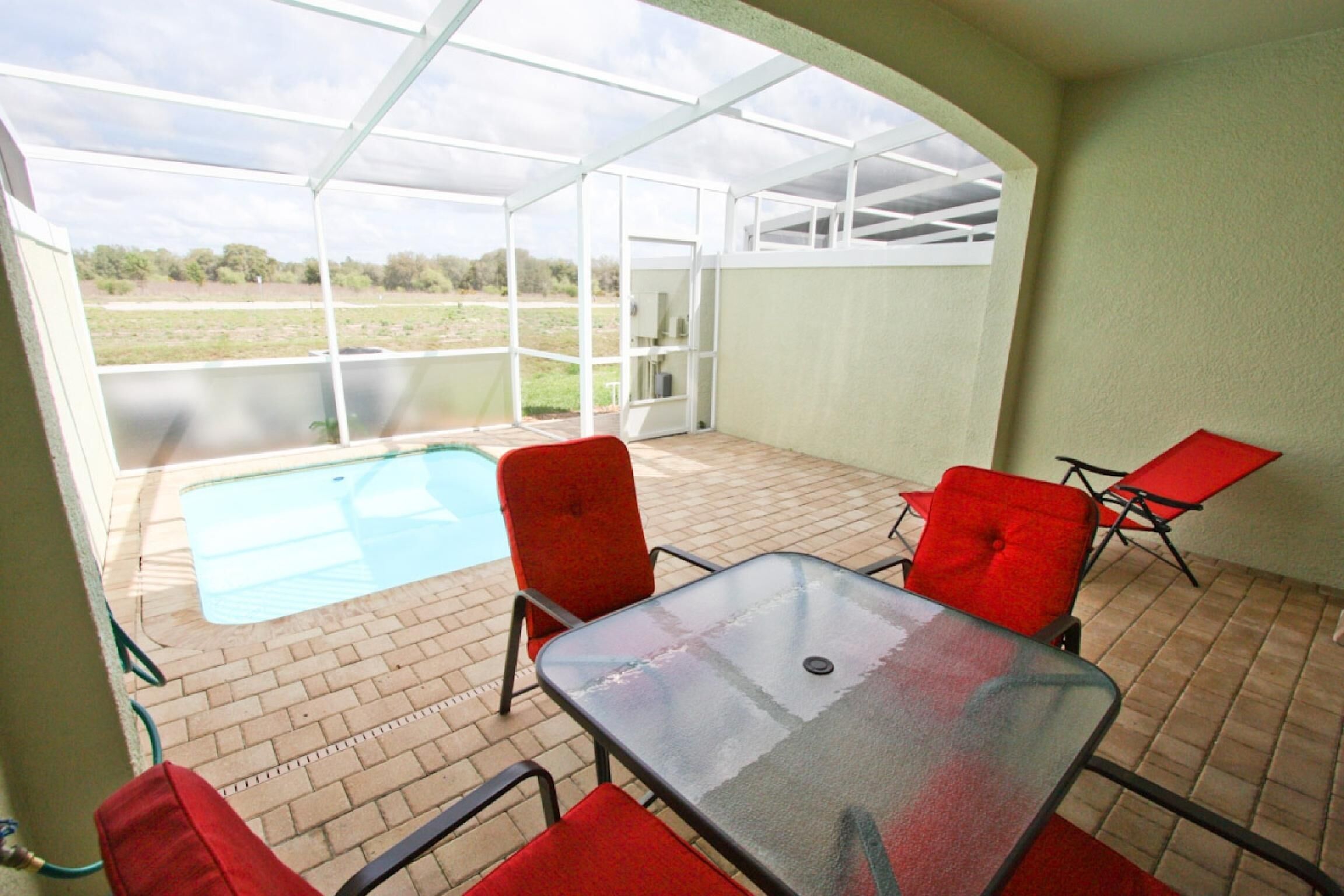 A covered patio with a glass table and red chairs, a swimming pool, and a view of a grassy field.