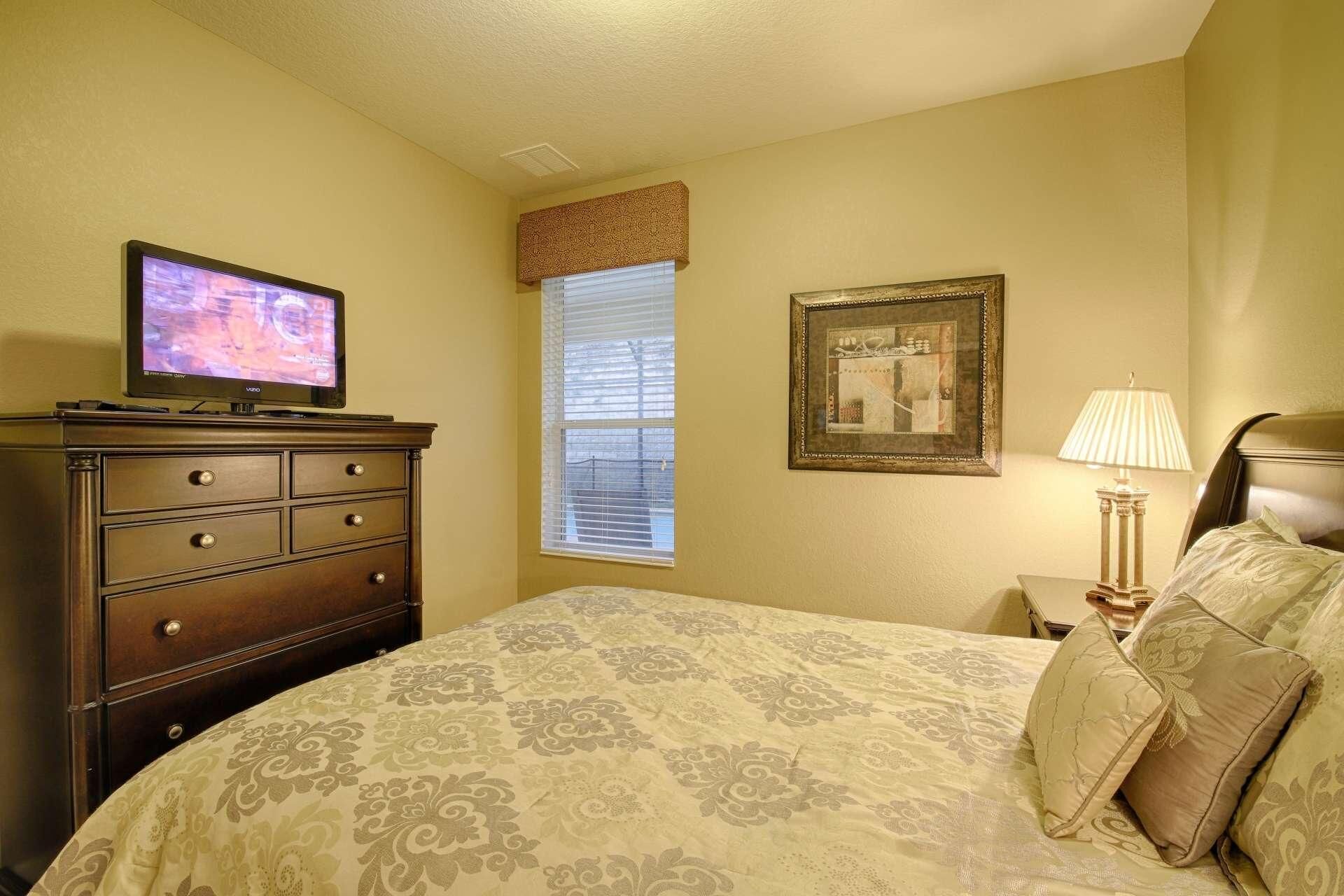 Een slaapkamer met een bed, een dressoir, een televisie, een raam met zonwering, een ingelijste foto en een bedlamp.