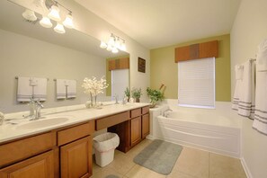 A modern bathroom with a large mirror, two sinks, a bathtub, and a shower. There are wooden cabinets, a trash bin, and a small rug on the floor. - 2540 Windsor Hills (Kissimmee)