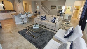 A modern living room with a sofa, coffee table, and bar stools. There is a kitchen area with a counter and appliances. A staircase is visible in the background. - 4201 Solterra (Davenport)
