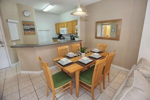 A dining area with a wooden table set for six, surrounded by green cushioned chairs. A kitchen with wooden cabinets and a white refrigerator is visible in the background. - 952 Lake Berkley (Kissimmee)