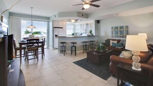 Una sala moderna con un sofá de cuero marrón, una mesa de centro y un televisor. Hay un comedor con una mesa y sillas, y una cocina con armarios blancos y una barra.