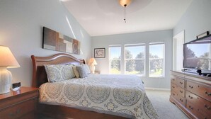 Ein Schlafzimmer mit einem Bett, Nachttischlampen, einer Kommode, einem Fernseher und großen Fenstern.