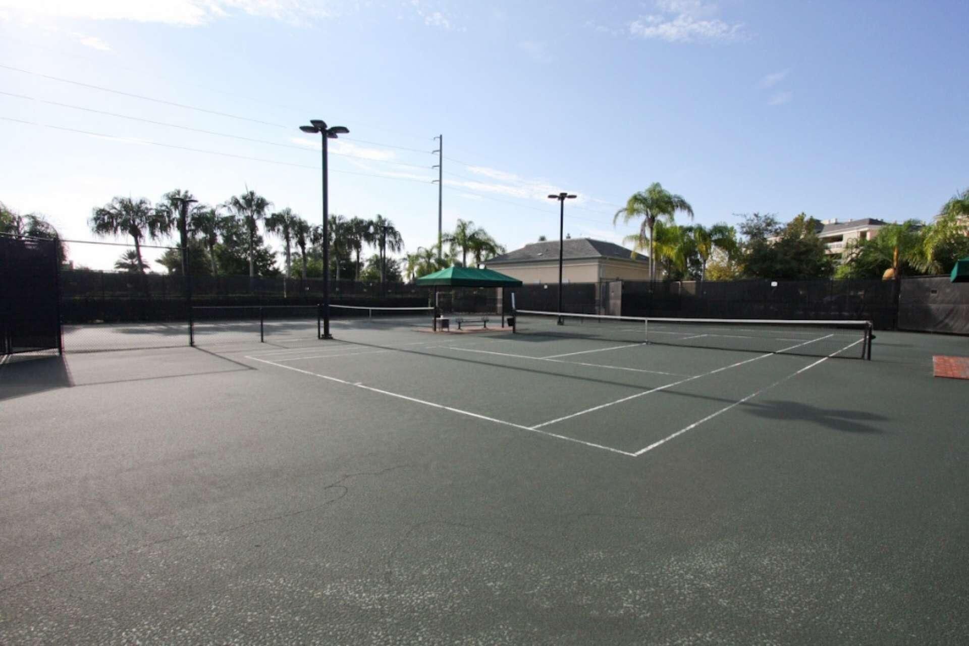 An outdoor tennis court with a green canopy shelter, surrounded by palm trees and a fence.