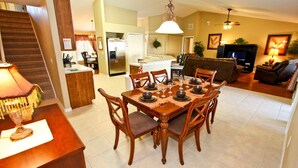 A dining area with a wooden table set for six, surrounded by chairs. There is a kitchen in the background with a refrigerator and a dining table. - 217 Legacy Park (Davenport)