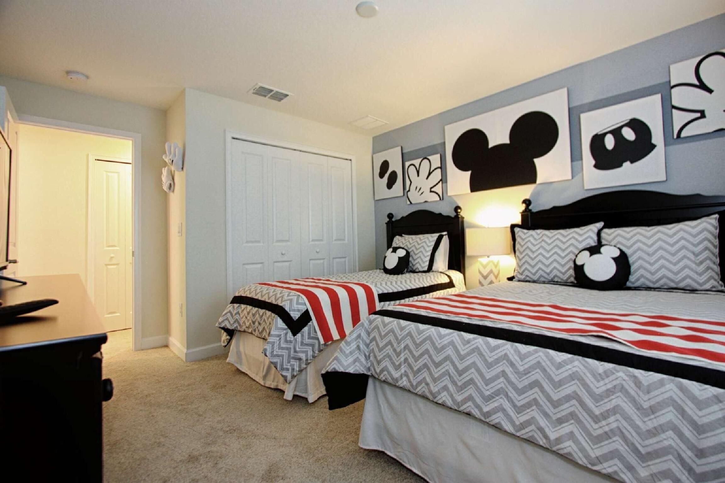 A bedroom with two beds, each with Mickey Mouse themed bedding. There are wall art pieces and a door visible.