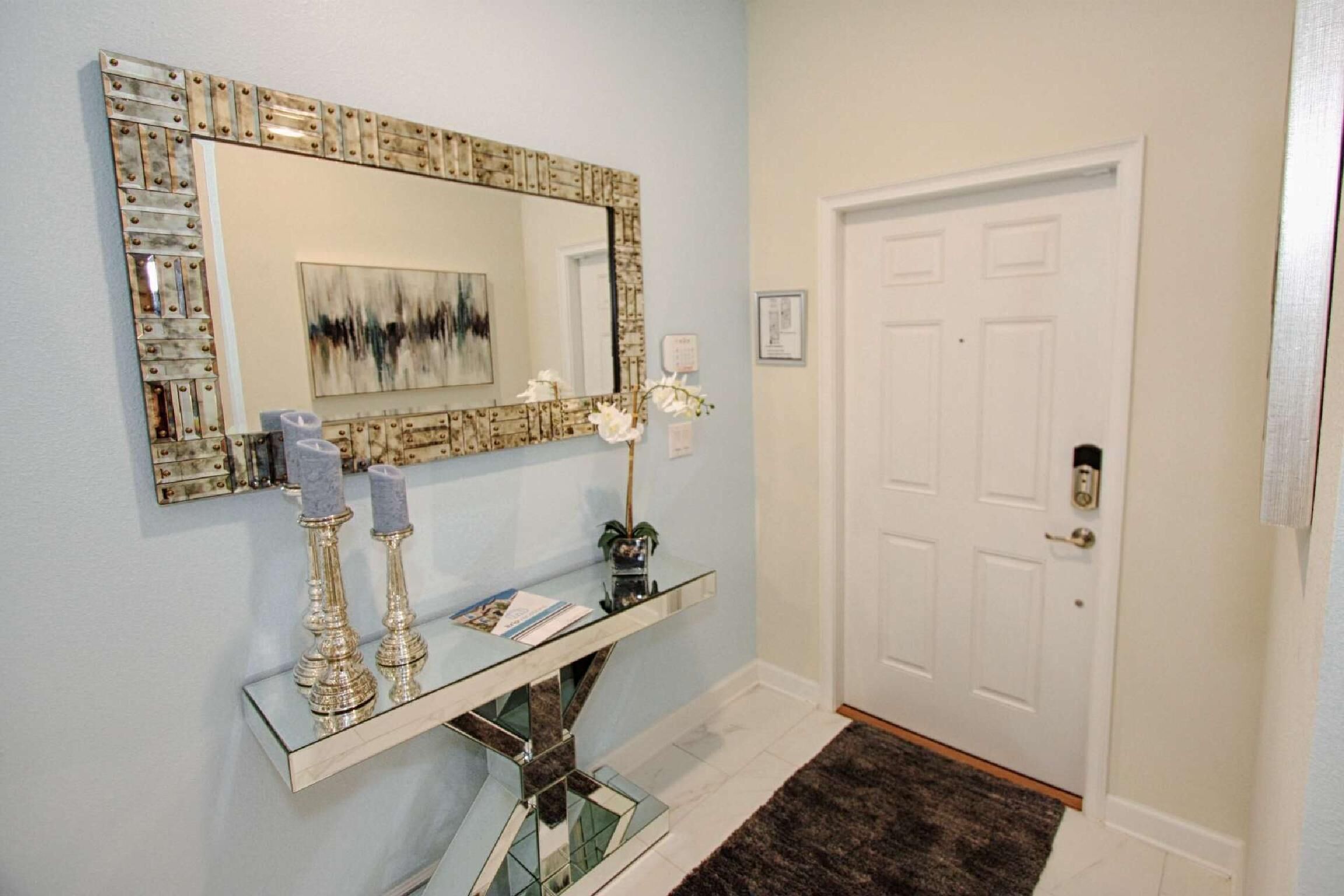 Un couloir avec une console ornée d’un miroir, une porte et un tableau abstrait encadré.