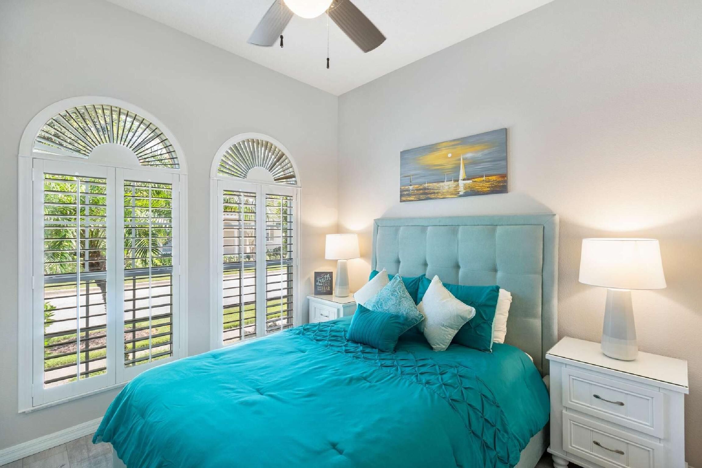 A bedroom with a large bed, two bedside tables, a ceiling fan, and a painting on the wall.