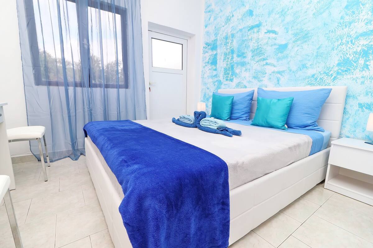 Un dormitorio con cama, mesita de noche, silla y ventana con cortinas traslúcidas.