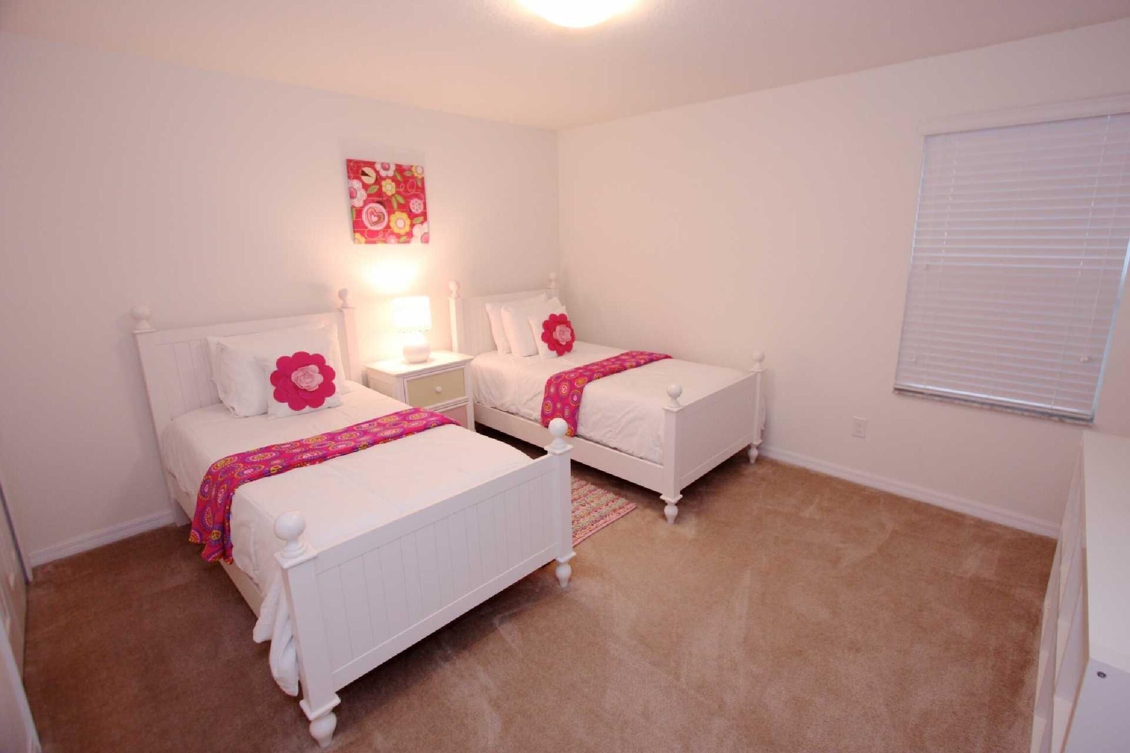 A bedroom with two white beds, a nightstand with a lamp, a floral artwork on the wall, and a window with blinds.