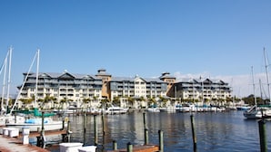 Una marina amb diverses embarcacions amarrades, un hotel al passeig marítim amb balcons, i un cel blau clar.