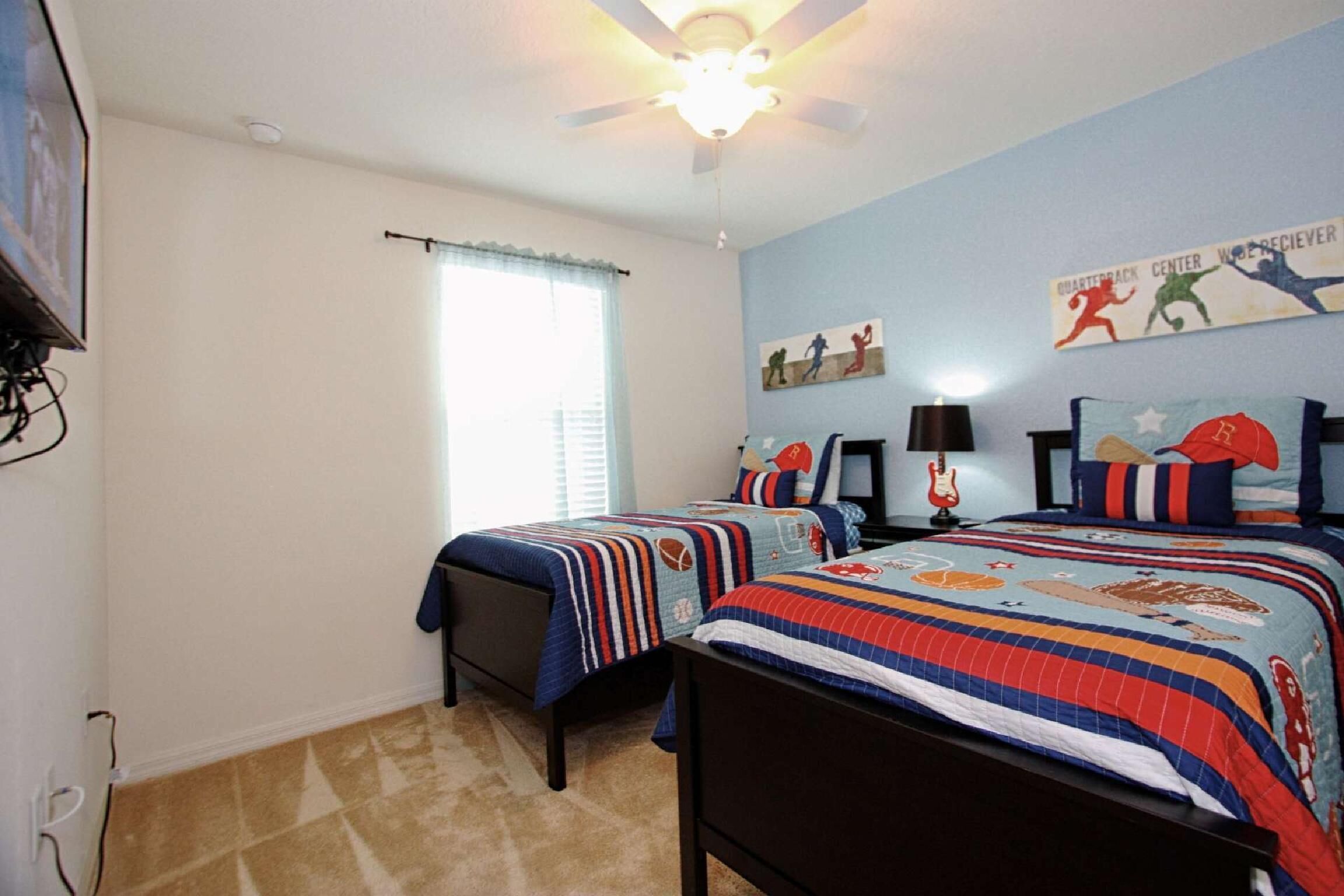 A bedroom with two beds, a ceiling fan, a window with curtains, and sports-themed wall art.