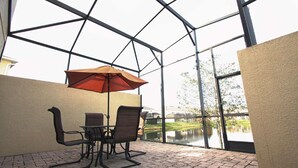 A covered patio with a table and chairs, a red umbrella, and a view of a lake through large windows. - 3207 Compass Bay (Kissimmee)