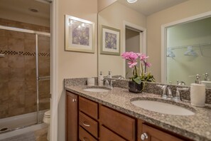 A bathroom with two sinks, a shower, and a toilet. There are framed pictures on the wall and a potted plant with pink flowers. - 2054 Windsor at Westside (Kissimmee)