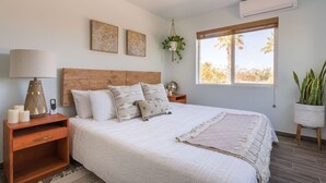 Un dormitorio con una cama grande, cabecera de madera, mesitas de noche, una planta colgante y una ventana con vistas a palmeras.