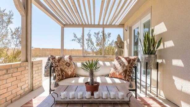 Un espacio de estar al aire libre techado con una mesa de madera, una planta en maceta y un banco con cojines estampados.
