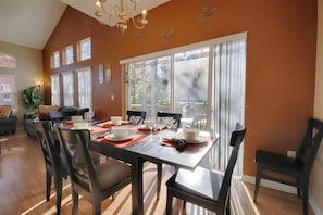 A dining area with a large table set for six, surrounded by chairs, with a view of a garden through large windows. - 8422 Emerald Island (Kissimmee)