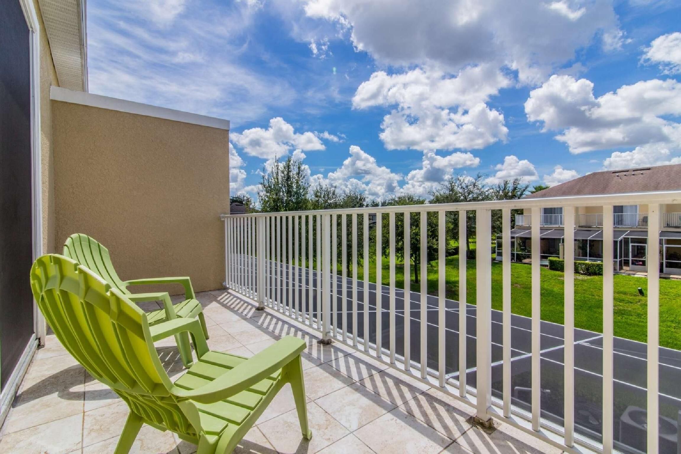A balcony with two green Adirondack chairs, a white metal railing, and a view of a parking lot and greenery.