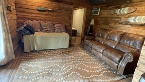 Interior acolhedor de uma cabana com piso de madeira, um tapete grande, um sofá de couro e uma cama com almofadas.