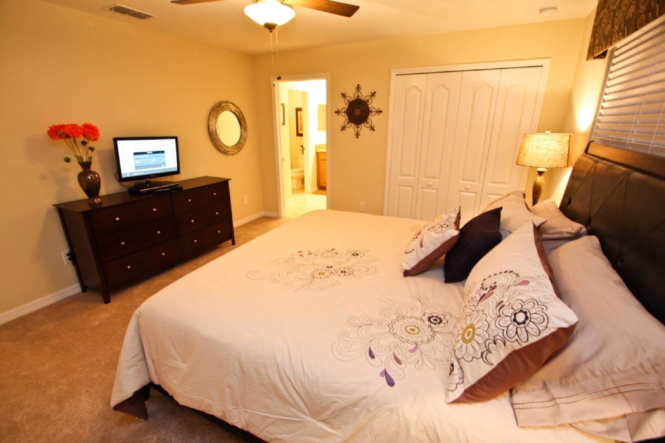 A bedroom with a large bed, a dresser, a TV, and a door leading to another room.