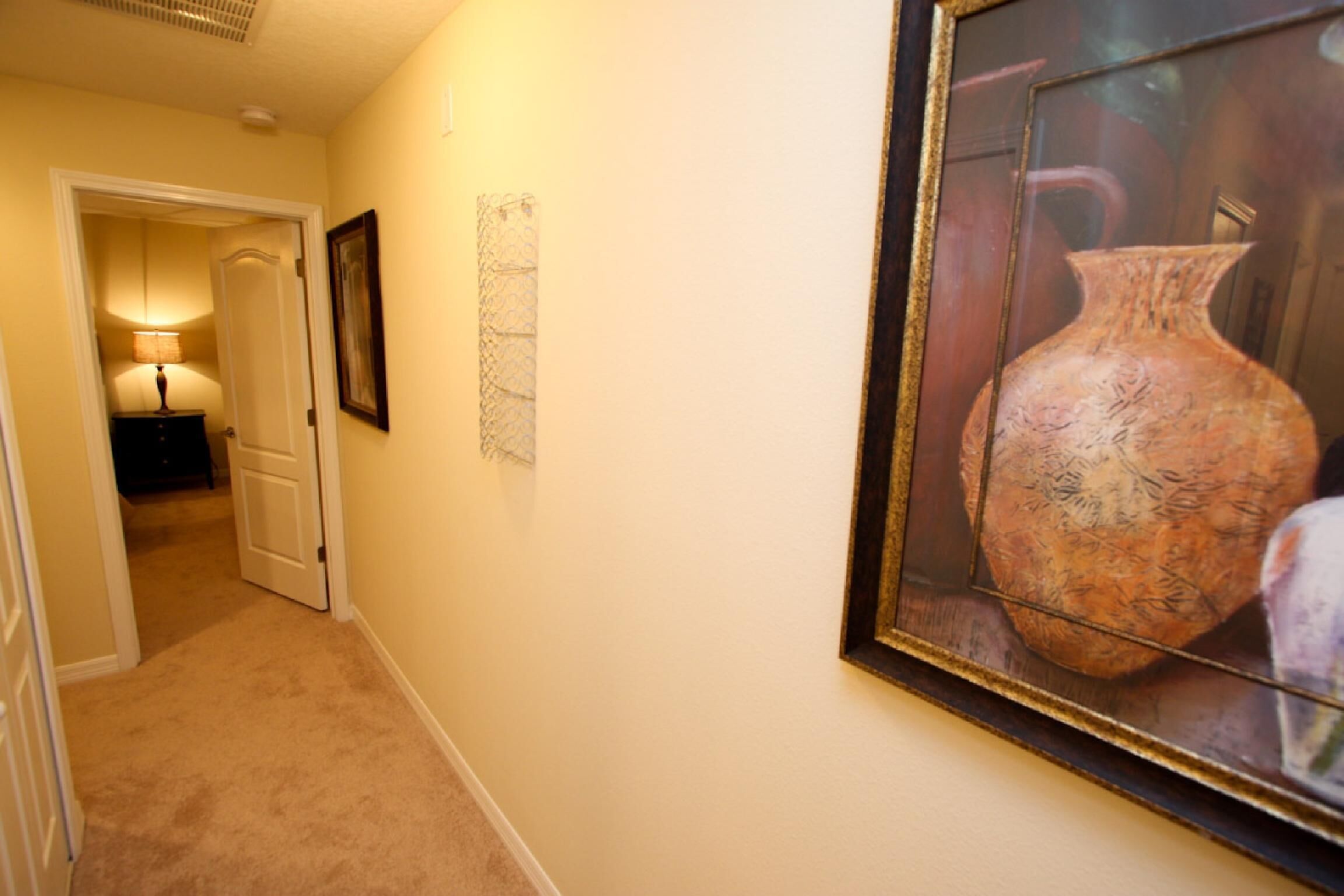 A hallway with a framed vase on the wall, a door leading to another room, and a lamp in the distance.