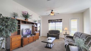 A living room with a TV, wooden entertainment unit, a ceiling fan, a sofa, a coffee table, and a window with blinds. - Vista Cay 4804 (Orlando)