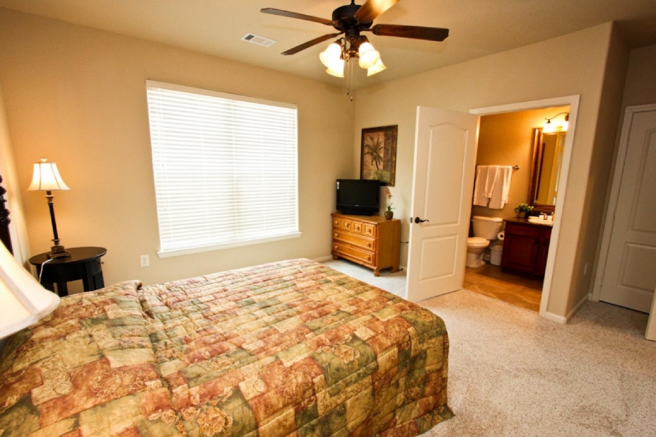 A bedroom with a bed, a ceiling fan, a dresser, a television, and a bathroom visible through an open door.