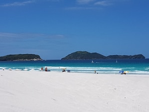 A beach with white sand, clear blue water, and distant green hills.