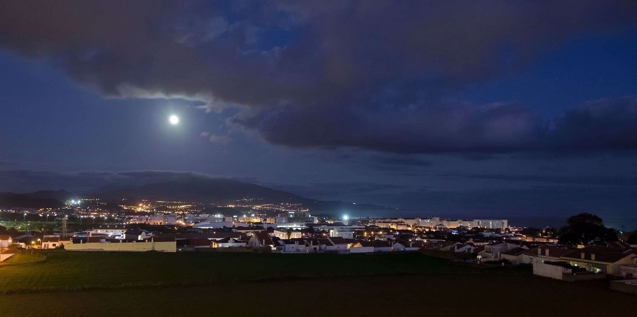 Ein Nachtblick auf eine Küstenstadt mit einem markanten Berg im Hintergrund, beleuchteten Gebäuden und einem sichtbaren Mond.