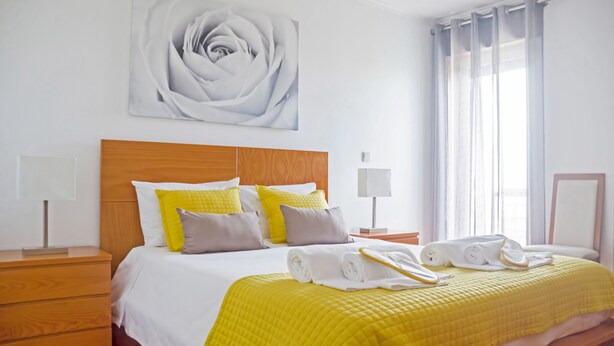 A neatly made bed with a yellow quilt, a wooden headboard, and a large wall-mounted rose painting.