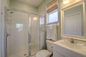 A bathroom with a glass-enclosed shower, a toilet, a sink with a mirror, and a window with a towel hanging on the wall.
