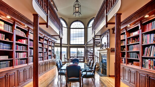 Une bibliothèque spacieuse avec des étagères en bois, une table centrale entourée de chaises et une grande fenêtre en arc de cercle.