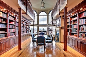 A spacious library with wooden bookshelves, a central table with chairs, and a large arched window. - Ghost Lake Manor House (Ghost Lake)