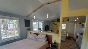 Quarto com cama, mesa de cabeceira e uma pequena escrivaninha com cadeira. Há uma janela com persianas, uma porta que dá para a cozinha e vista para o exterior.