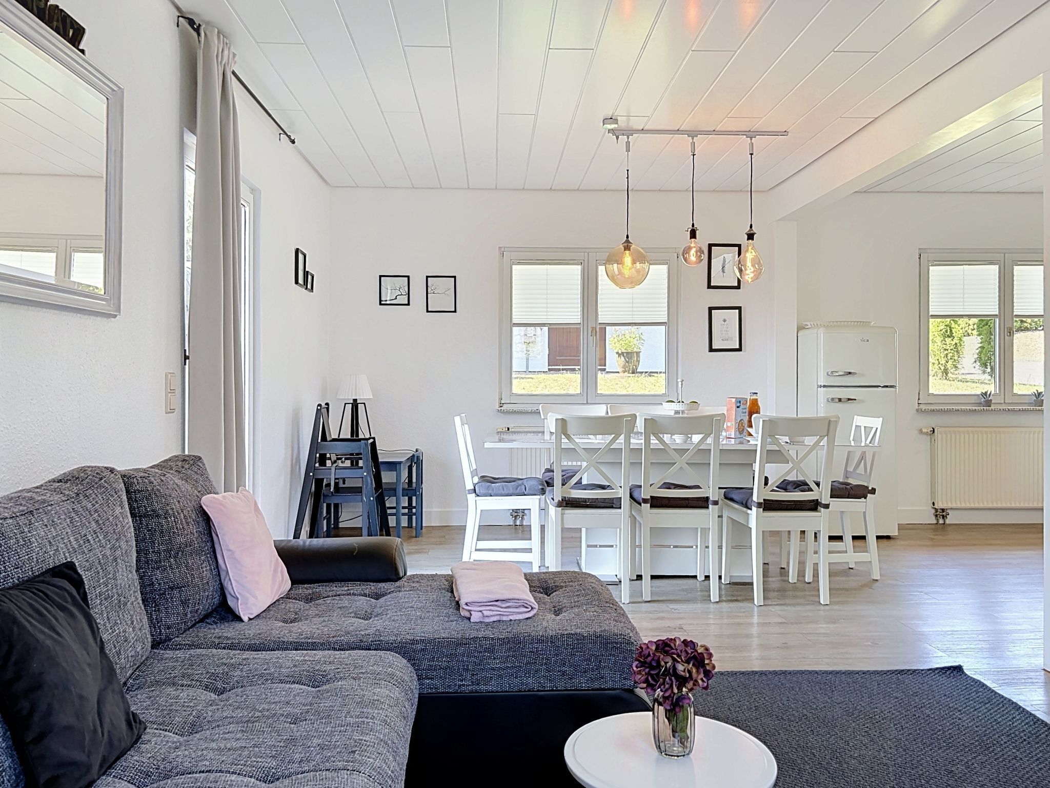 A spacious living room with a grey sofa, a dining area with white chairs, and a kitchen with a white refrigerator.