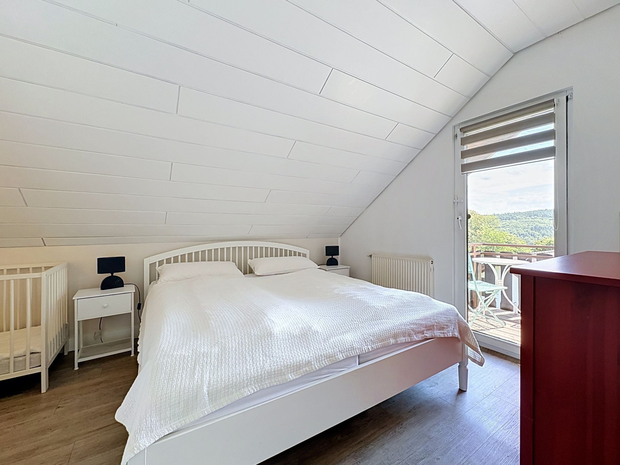 A bedroom with a sloped ceiling, a bed, a nightstand, a lamp, a radiator, and a view through a window.