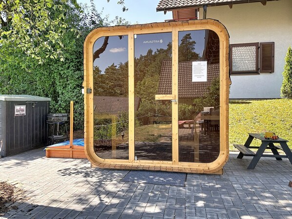 A wooden structure with a glass door, a patio area with a table and chairs, and a garden with a hedge.