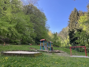 Un terrain de jeux avec un toboggan et des balançoires, entouré d’arbres et d’herbe.