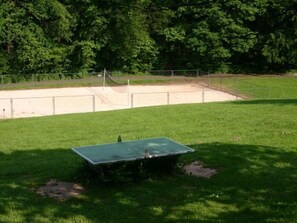 Une table de ping-pong verte se trouve dans un espace herbeux, avec un court de tennis entouré de clôtures en arrière-plan.