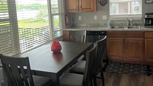A kitchen with a dining table, chairs, wooden cabinets, a window with blinds, and a view of greenery outside.
