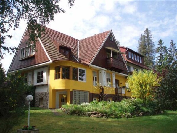 Una casa de dos pisos con fachada amarilla y techo marrón, rodeada de follaje y árboles.