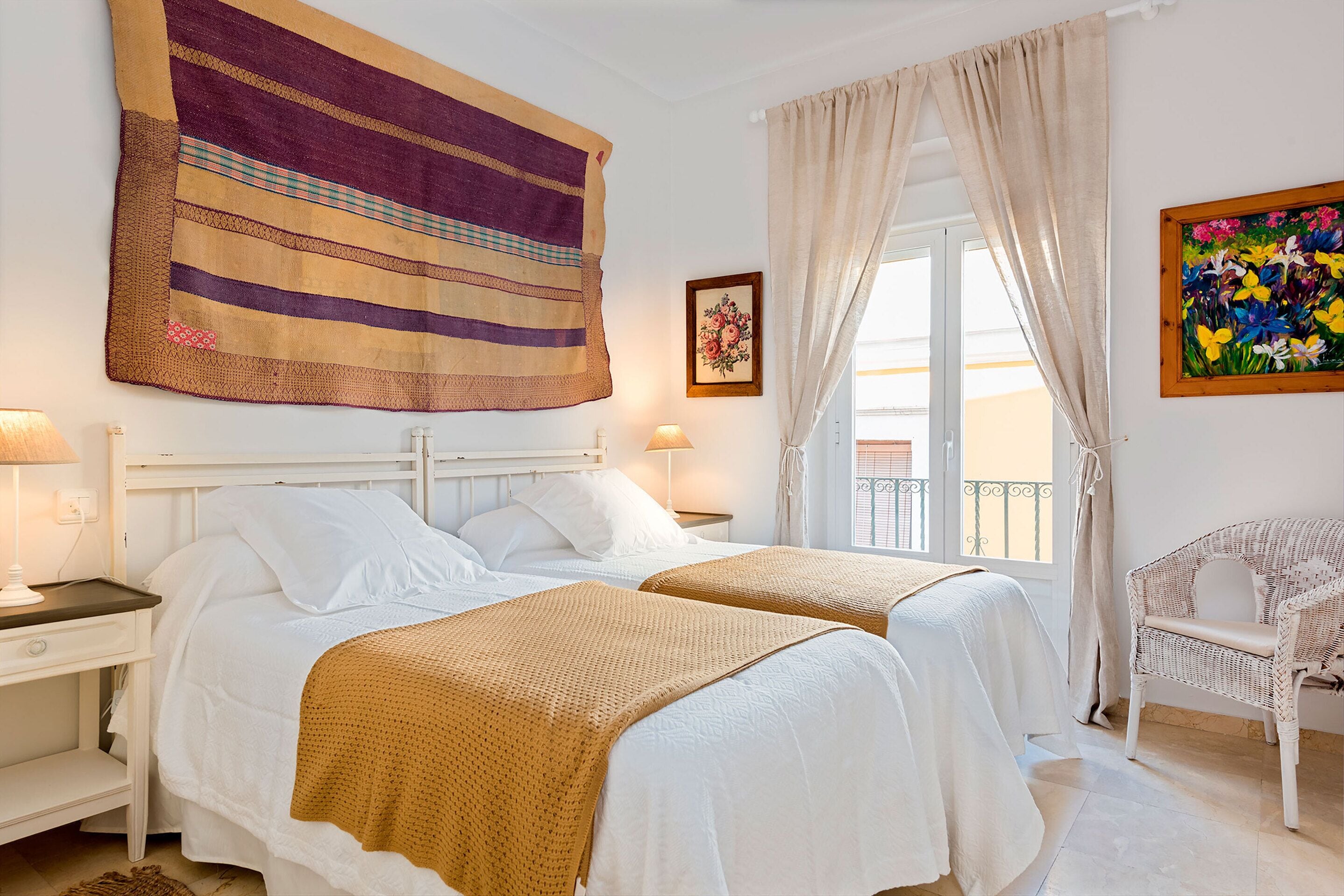 A hotel room with two beds, a large tapestry, a chair, and a balcony with a railing.
