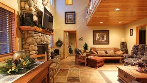 A cozy living room with a stone fireplace, a wooden ceiling, and a large window with blinds. - Walk 2 Ski/Hiking/Biking Trails w/Spa/Pool Table/Sauna/Fenced Yard, Pet Friendly (Big Bear Lake)