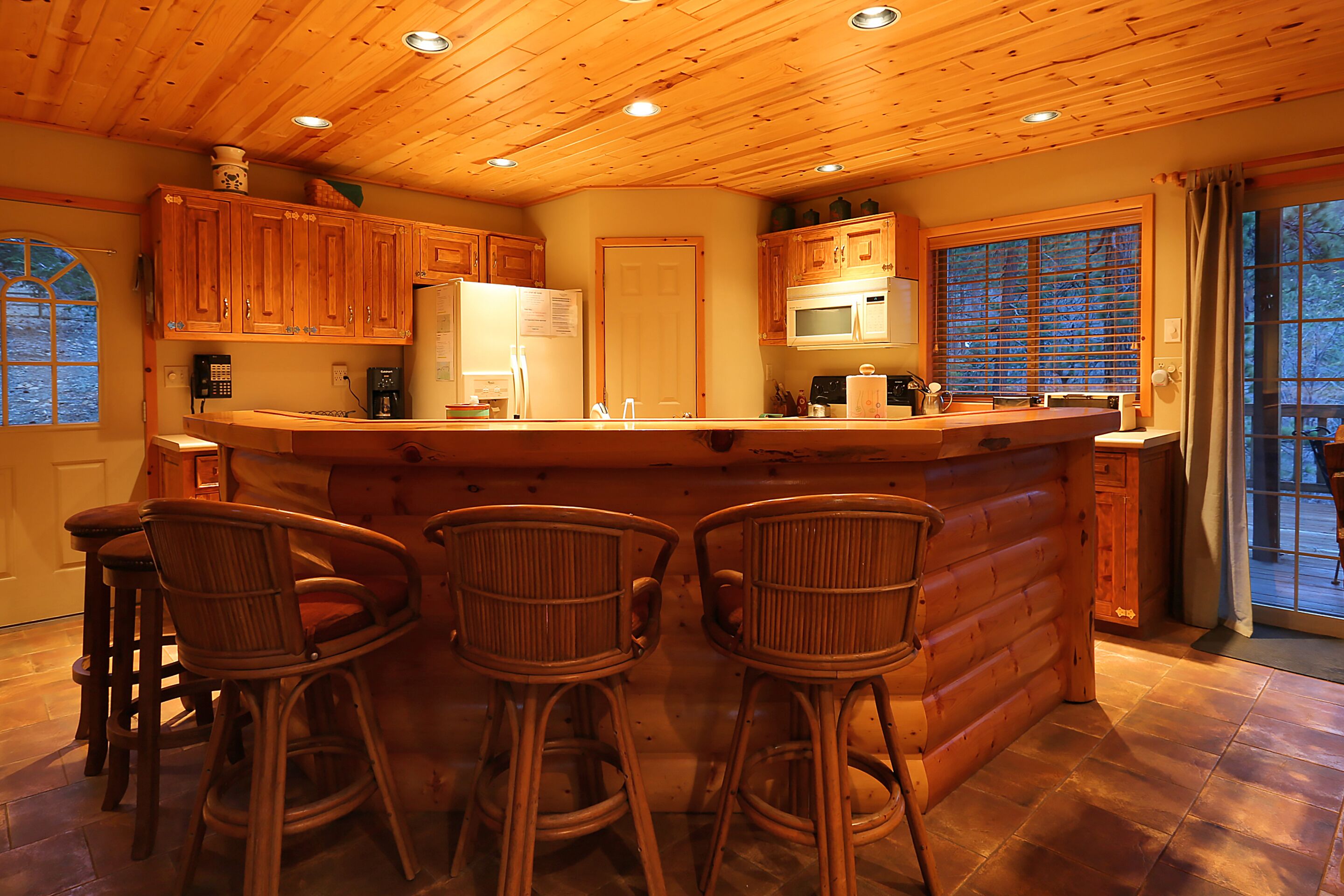 Cocina de cabaña de madera con armarios de madera, una isla central con taburetes de bar, un refrigerador, microondas y una ventana con persianas.