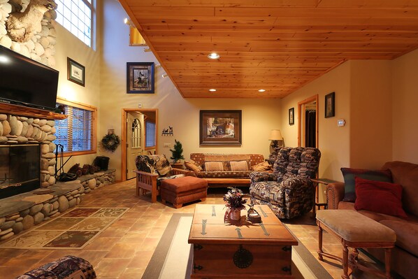 A cozy living room with a stone fireplace, wooden ceiling, and comfortable seating including a sofa, armchair, and ottoman.