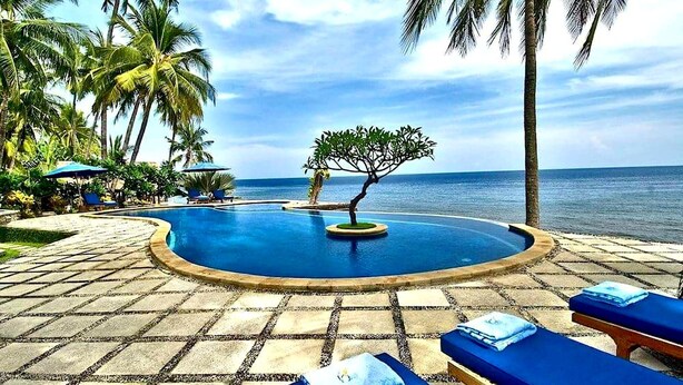 A poolside area with lounge chairs and umbrellas, surrounded by palm trees and a view of the ocean.