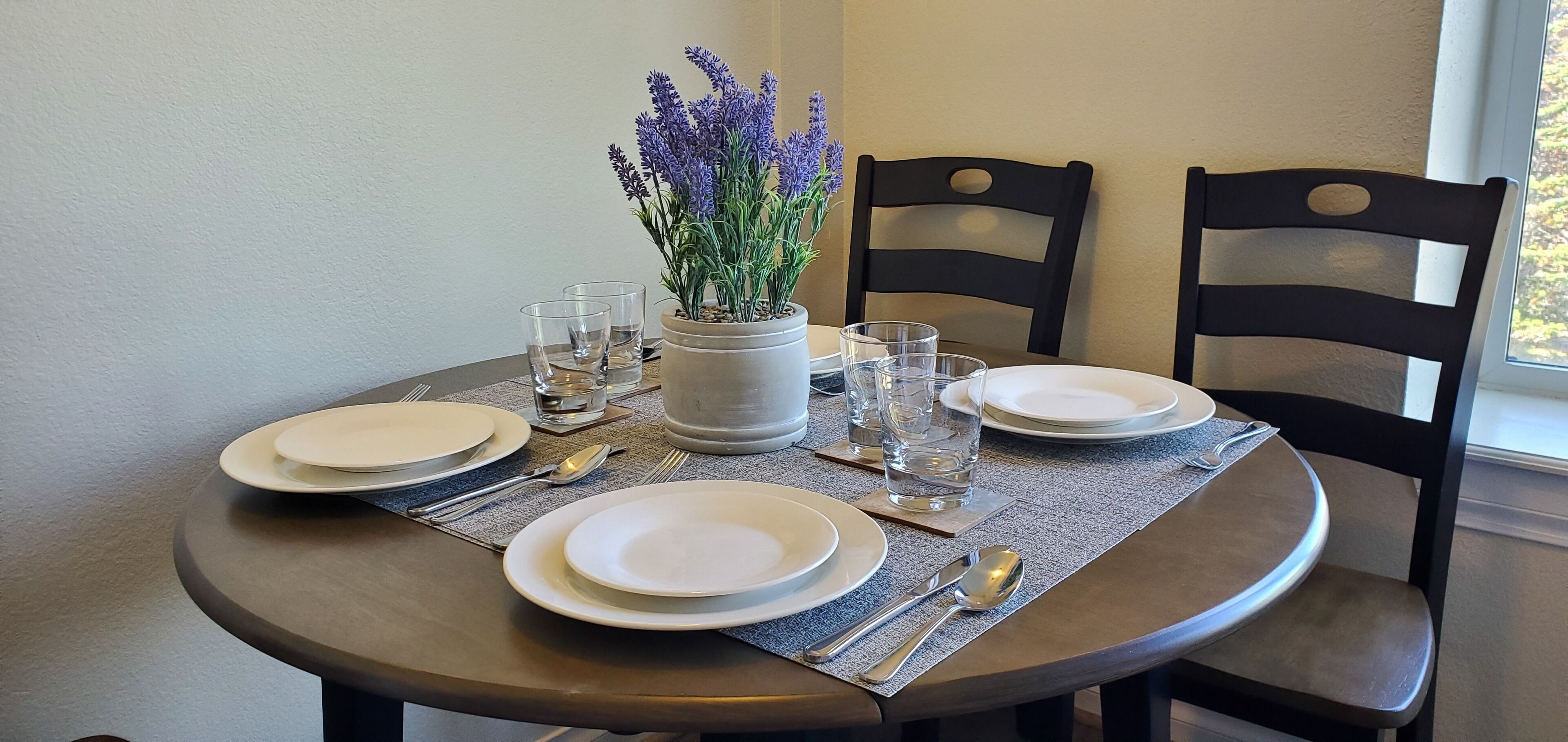 A round dining table set with white plates, clear glasses, and silverware, accompanied by a potted lavender plant.